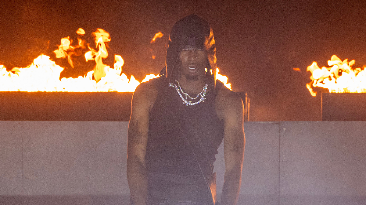 Playboi Carti performs on stage wearing a black tank top and black head covering, with fire blazing in the background