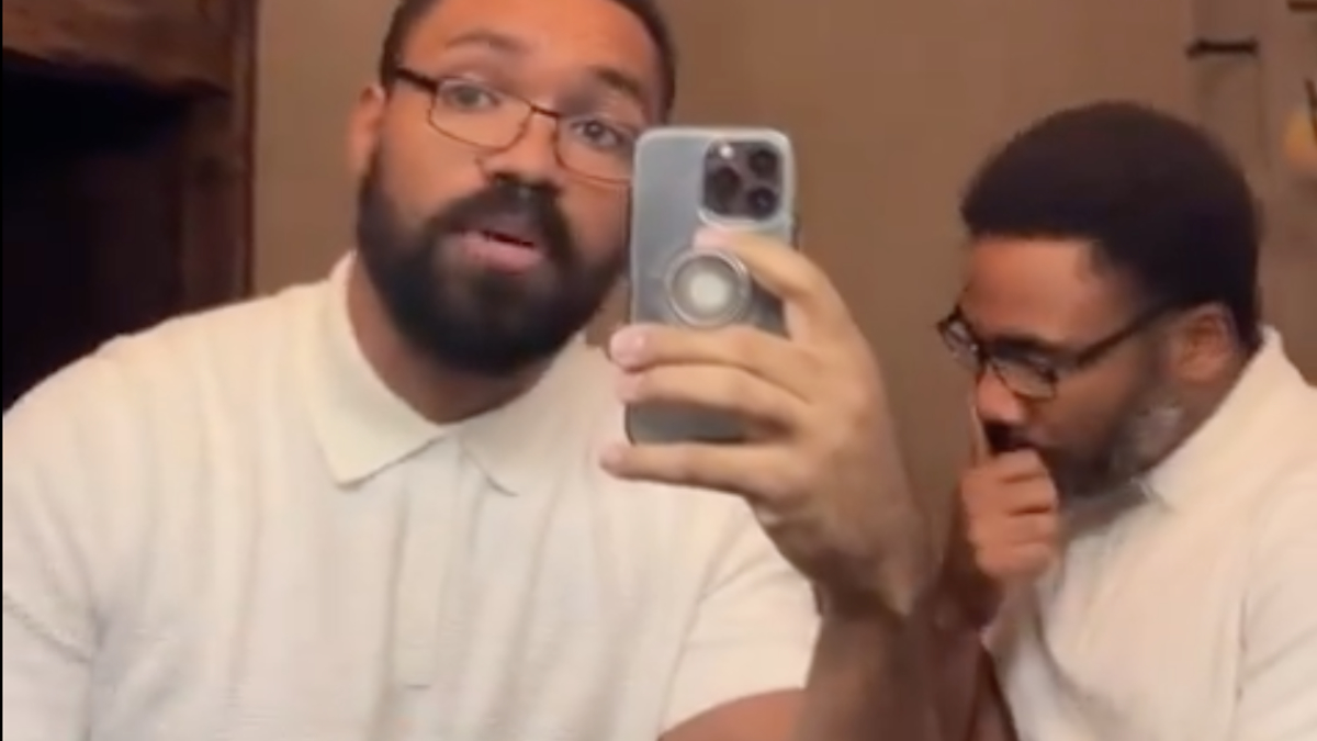 Two men are in a bathroom. One man wearing glasses is taking a selfie, while the other is standing behind him, deep in thought with his hand on his chin
