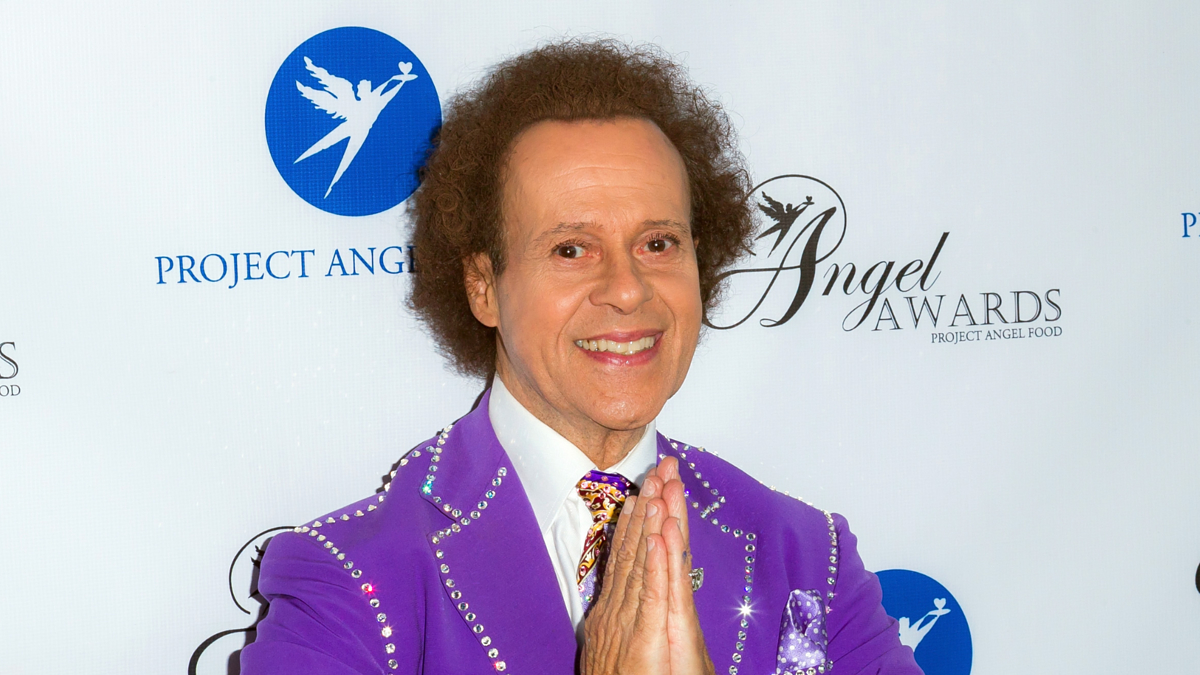 Richard Simmons, wearing a sparkling purple jacket, poses with hands clasped at the Project Angel Food Angel Awards event backdrop