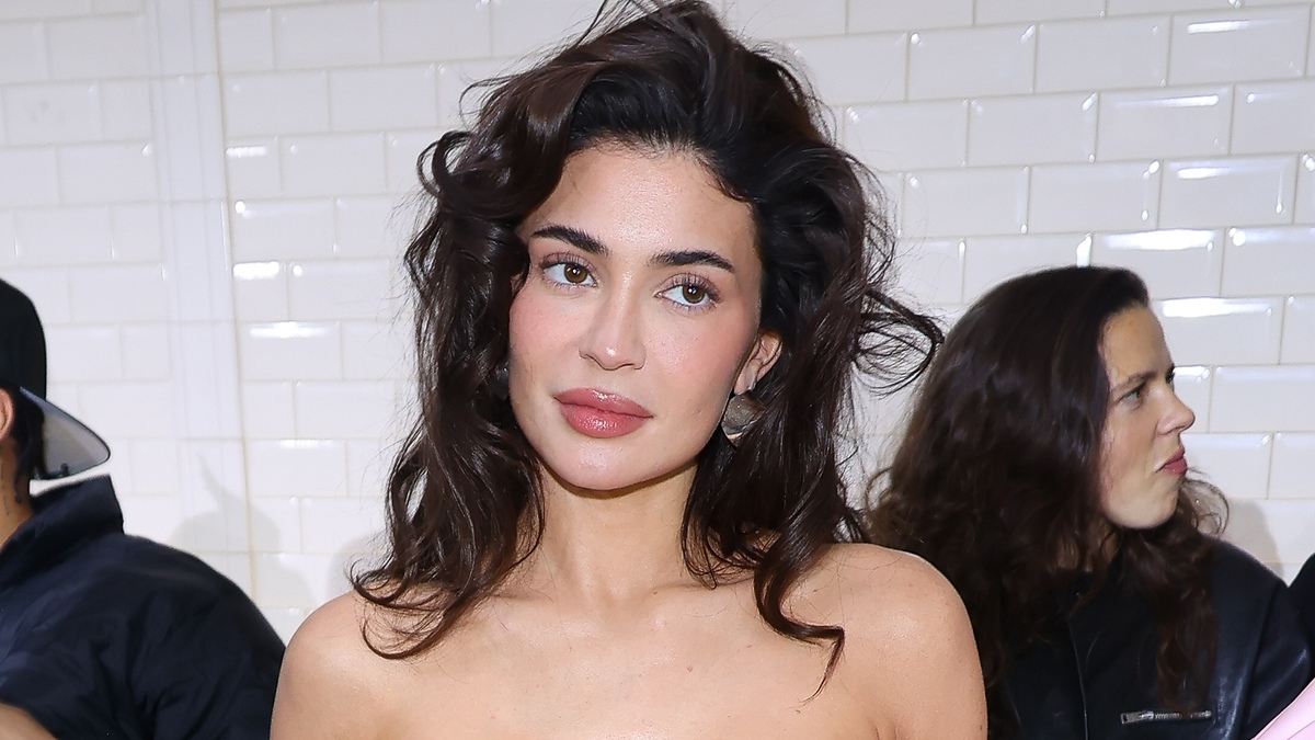A woman with wavy hair and bare shoulders poses, with a tiled wall background and another person partially visible behind her. Names are unknown