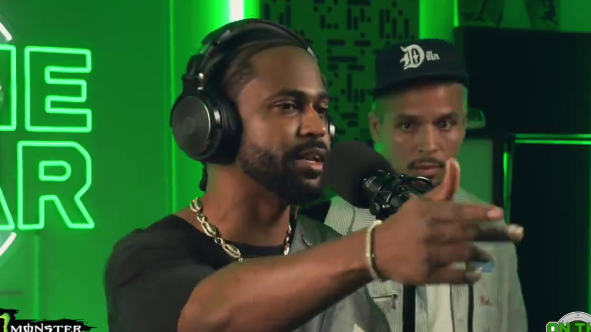 Big Sean performs into a microphone in a studio, wearing headphones and a black t-shirt with a chain necklace. Another man stands behind him