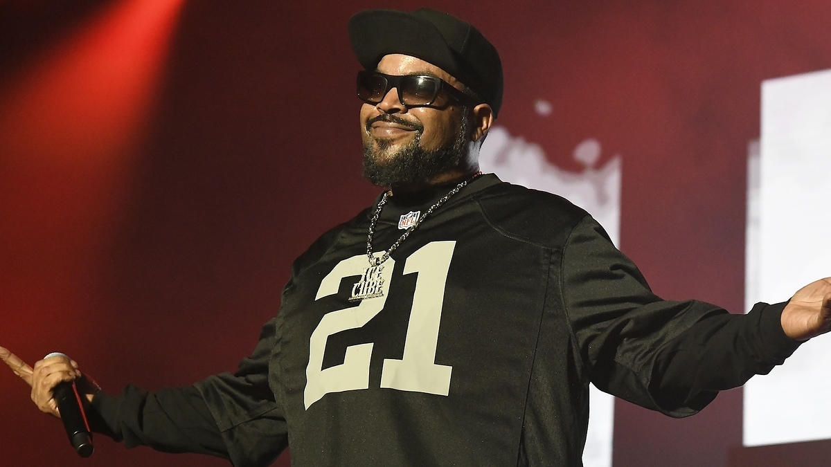 Ice Cube performs on stage, wearing sunglasses, a black cap, and a black jersey with the number 21
