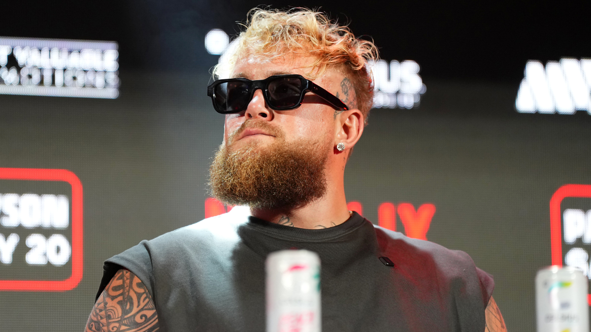 Jake Paul, wearing sunglasses and a sleeveless top, poses in front of a backdrop with logo text at an event