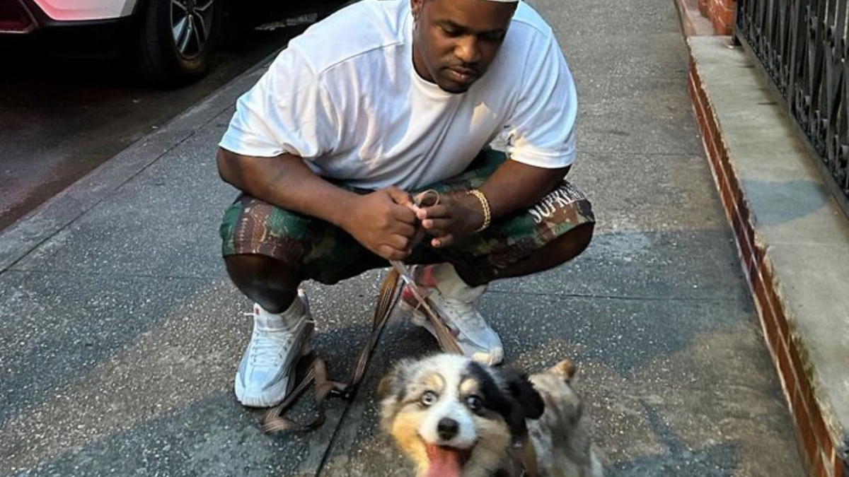 A$AP Ferg crouches on a sidewalk, holding a leash attached to his Australian Shepherd, who has a happy expression. A$AP Ferg wears a white t-shirt and camo shorts