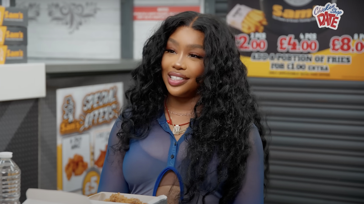 SZA sits in a restaurant, smiling while wearing a blue sheer top. The background shows menu prices and meal options displayed on the wall
