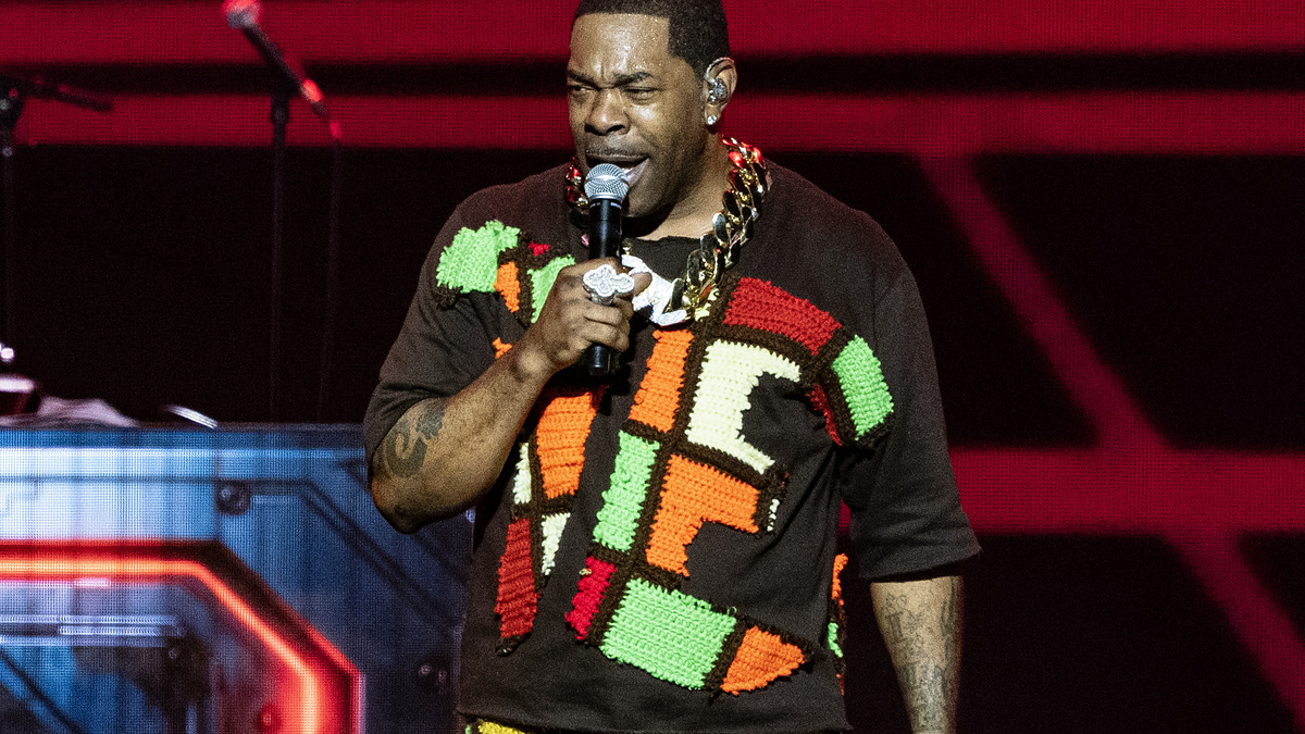 Busta Rhymes performs on stage in a colorful, patchwork outfit. A DJ stands in the background next to a digital display of Busta Rhymes' name