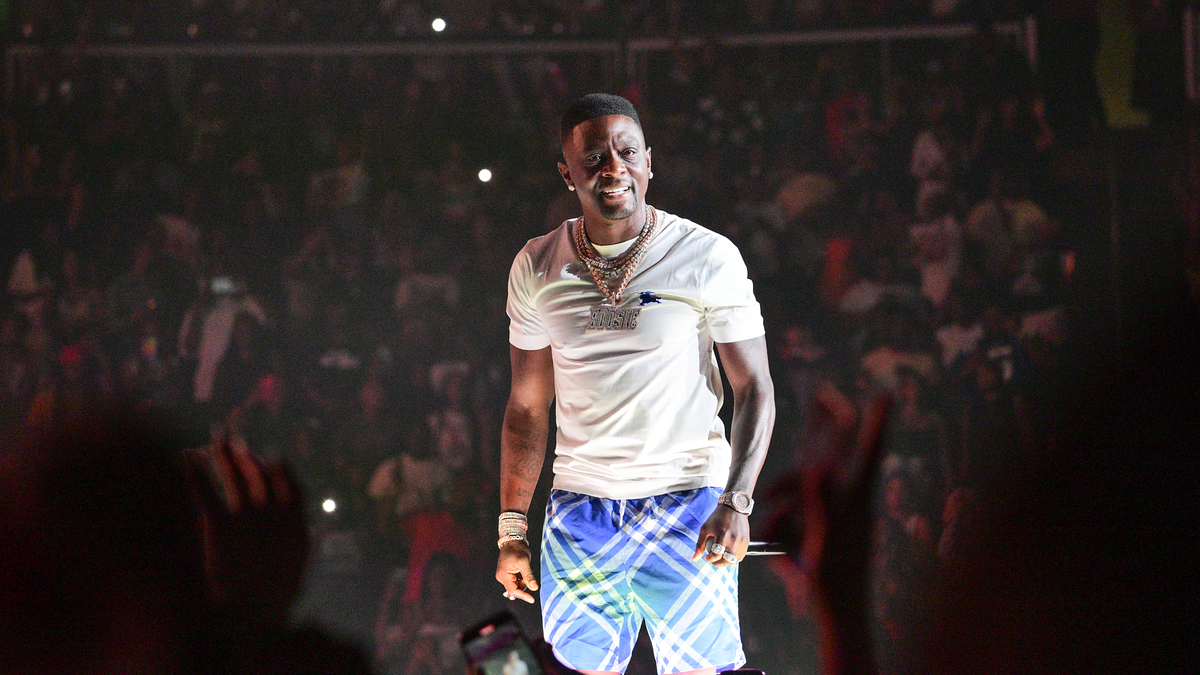Boosie Badazz on stage performing in front of a cheering crowd, wearing a white t-shirt, gold chain necklaces, and patterned shorts