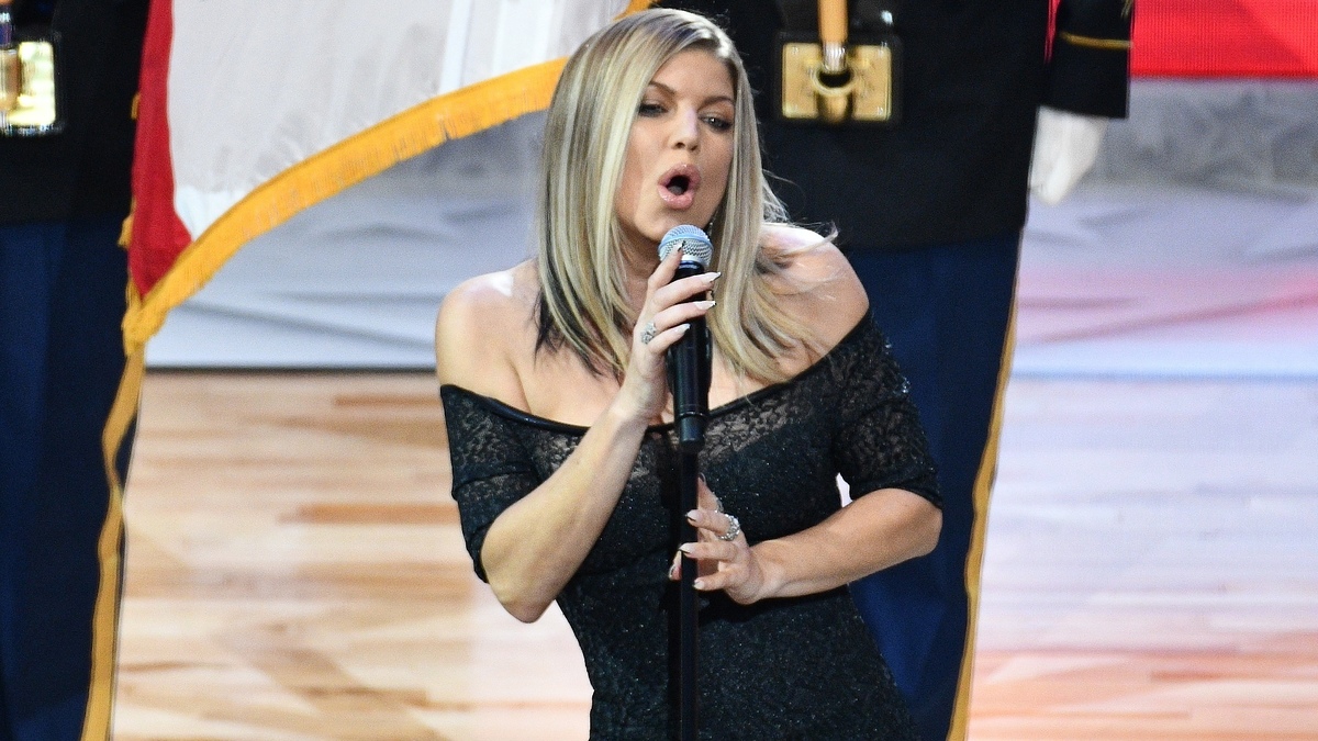Fergie sings into a microphone while wearing an off-shoulder dress, performing in front of military personnel holding a flag