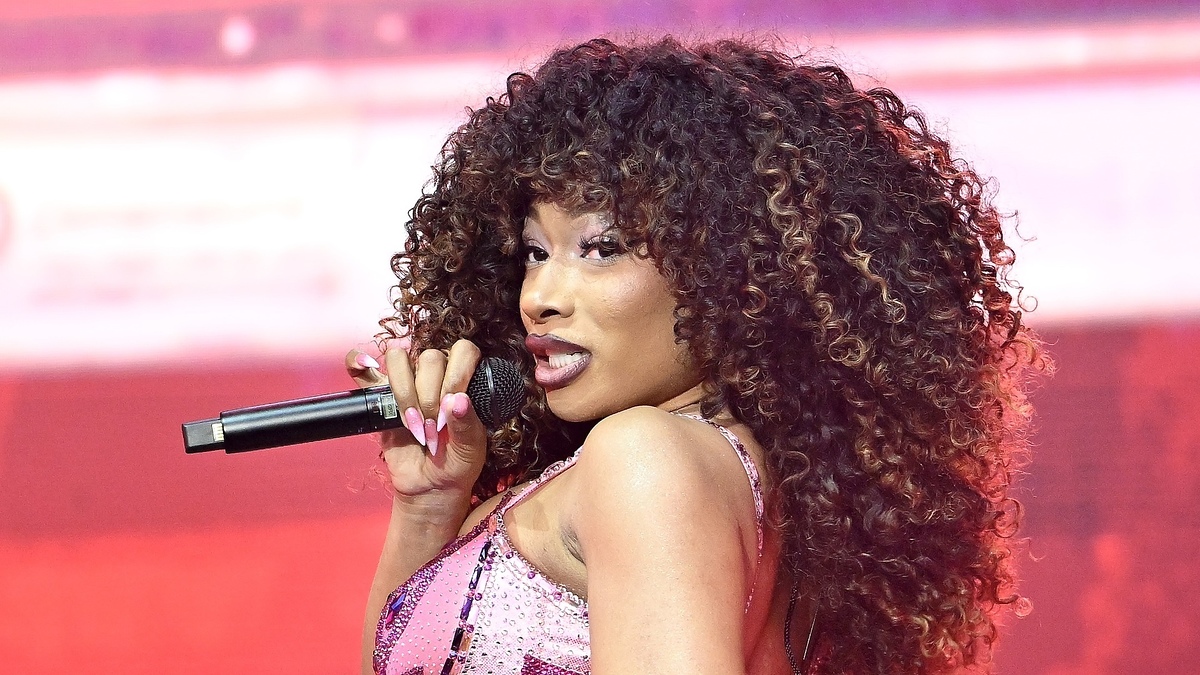 Megan Thee Stallion performs on stage, holding a microphone, with voluminous curly hair and wearing a sparkling outfit