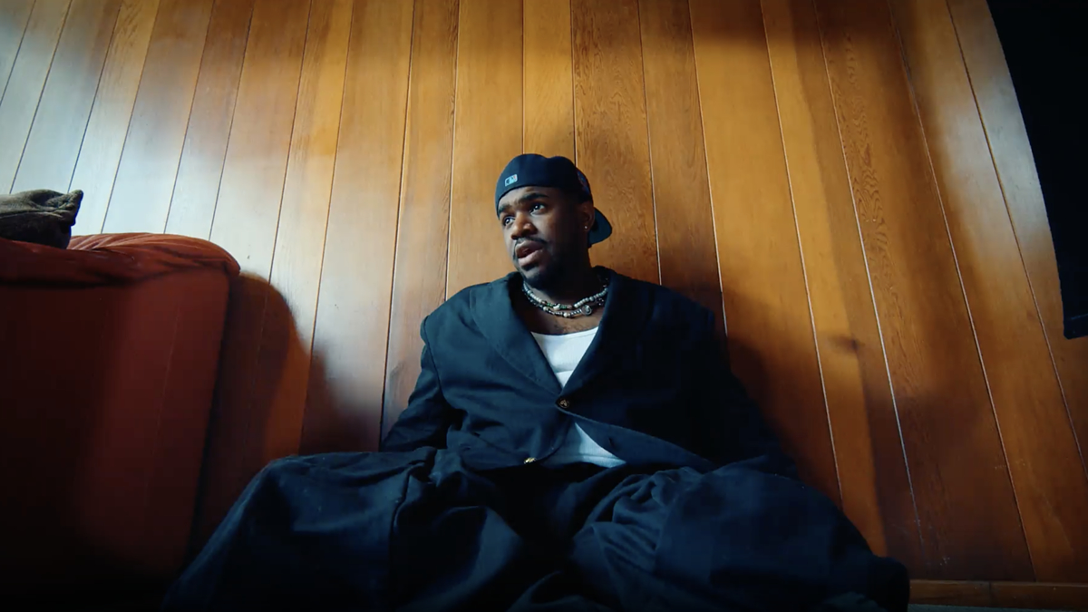 A person in black oversized clothing and a backwards cap sits against a wooden wall, next to a red sofa, looking into the distance