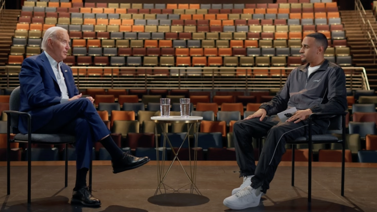 Joe Biden and Nate Burleson seated and conversing in an empty auditorium during an interview. Biden wears a suit, Burleson wears a tracksuit and sneakers