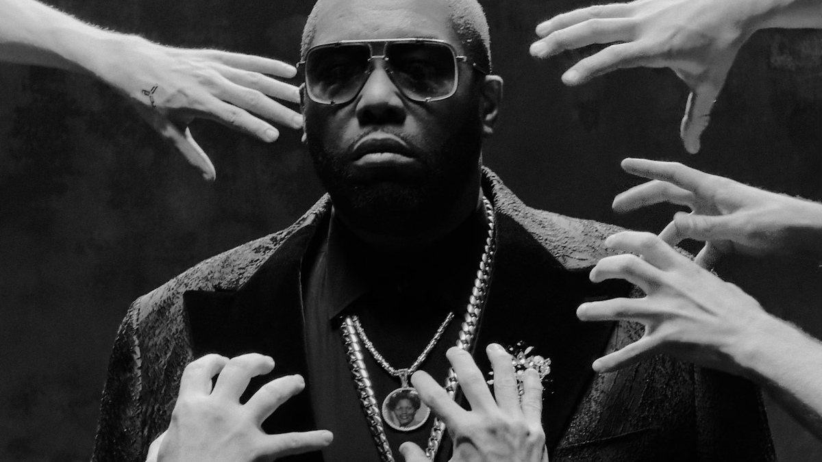 A man with sunglasses wears a dark jacket and multiple necklaces, surrounded by several outstretched hands reaching towards him