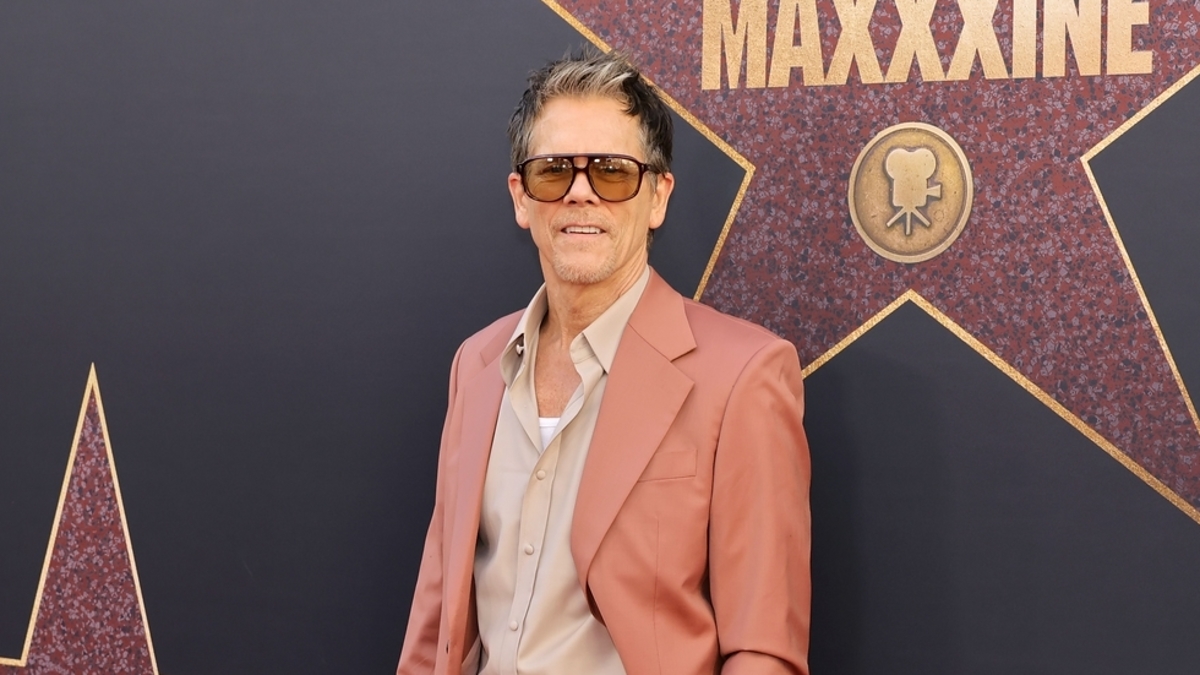 Man in a stylish suit and sunglasses posing in front of a wall with star decorations and the word "MAXXXINE."