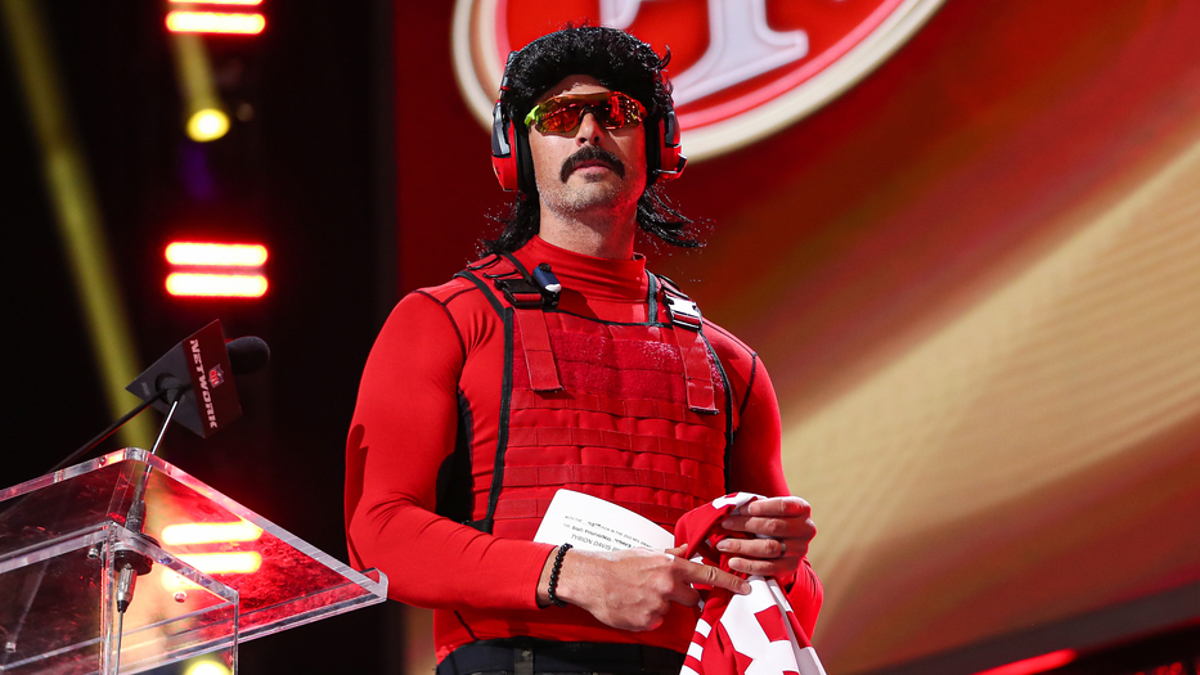 Dr. DisRespect stands on stage holding a San Francisco 49ers jersey, wearing a tactical red vest, sunglasses, headphones, and a wig with a mustache