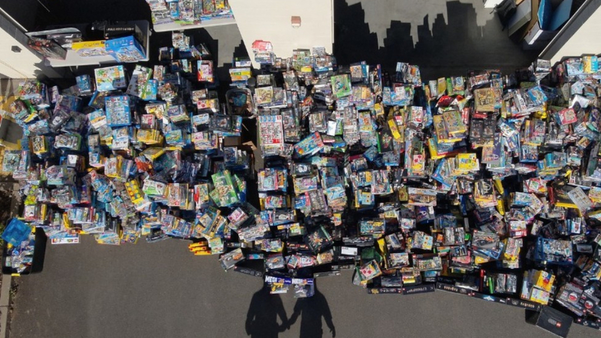 A large assortment of various toy packages are spread out on the ground, seemingly in a chaotic manner. Two shadows of people are visible in the foreground