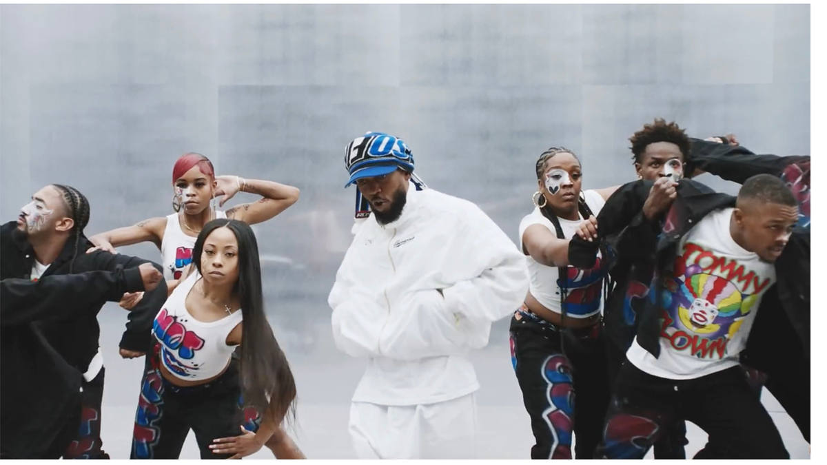 Kendrick Lamar in a white tracksuit and blue hat dances with a group of dancers in matching outfits, featuring graphic designs and face makeup