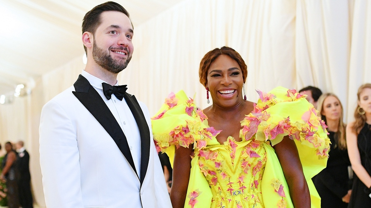 Alexis Ohanian and Serena Williams on a red carpet. Ohanian wears a black and white tuxedo; Williams in a vibrant dress with ruffled shoulders and floral designs