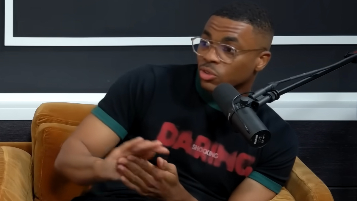 Vince Staples, wearing glasses and a T-shirt, speaking into a microphone while seated on a couch during an interview