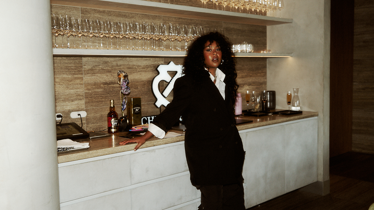 SZA stands in a chic, modern room with shelves of glasses and a champagne sign in the background. She wears a stylish, oversized coat