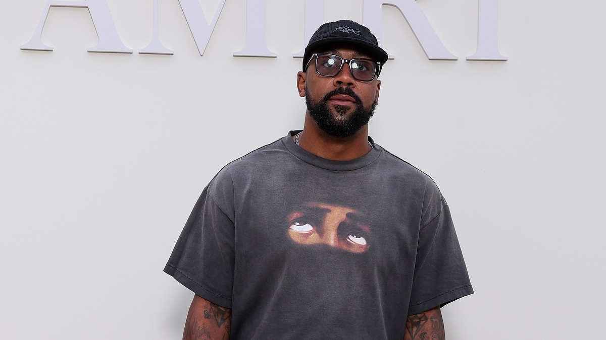 A man stands in front of an "AMIRI" sign wearing glasses, a black cap, and a T-shirt featuring an image of eyes looking upward