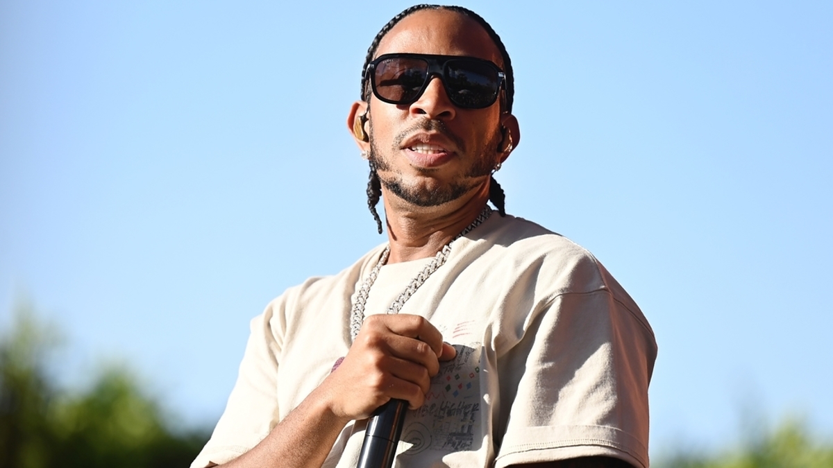 Ludacris is on stage wearing sunglasses, a beige T-shirt, and a silver chain, holding a microphone