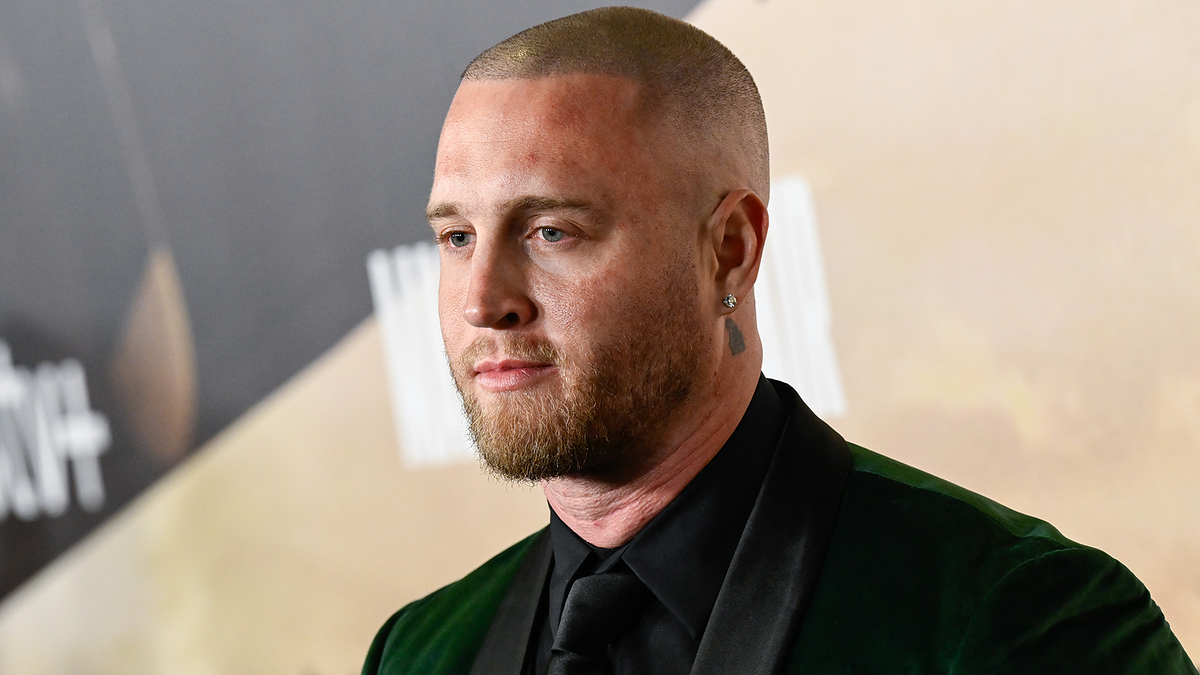 Man in a green velvet suit and black shirt stands on a red carpet at an event, posing for photos