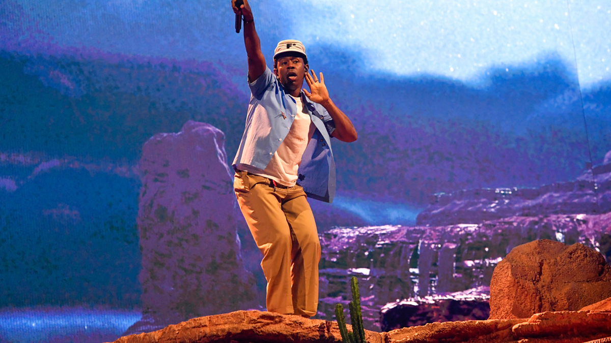 Tyler, the Creator performs on stage, wearing a blue hat, blue shirt, and beige pants, with a backdrop resembling a desert landscape