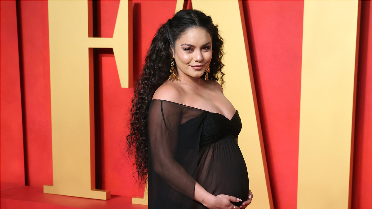 Vanessa Hudgens, posing on a red carpet in an elegant, off-the-shoulder black dress with sheer sleeves, cradles her baby bump. She wears gold earrings