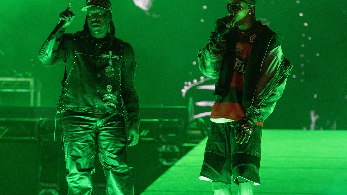 Two performers in stylized outfits are on a green-lit stage during a music event