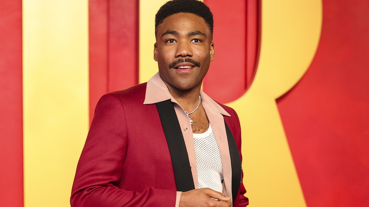 Donald Glover poses on the red carpet in a stylish suit jacket with a pink collar and black accents, paired with a mesh shirt underneath