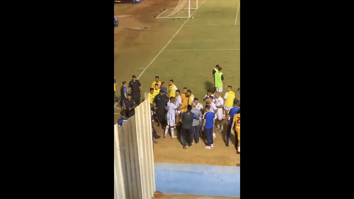 A group of soccer players and referees gather on the field, engaging in what appears to be a heated discussion near the goal. The scene is on a sports field