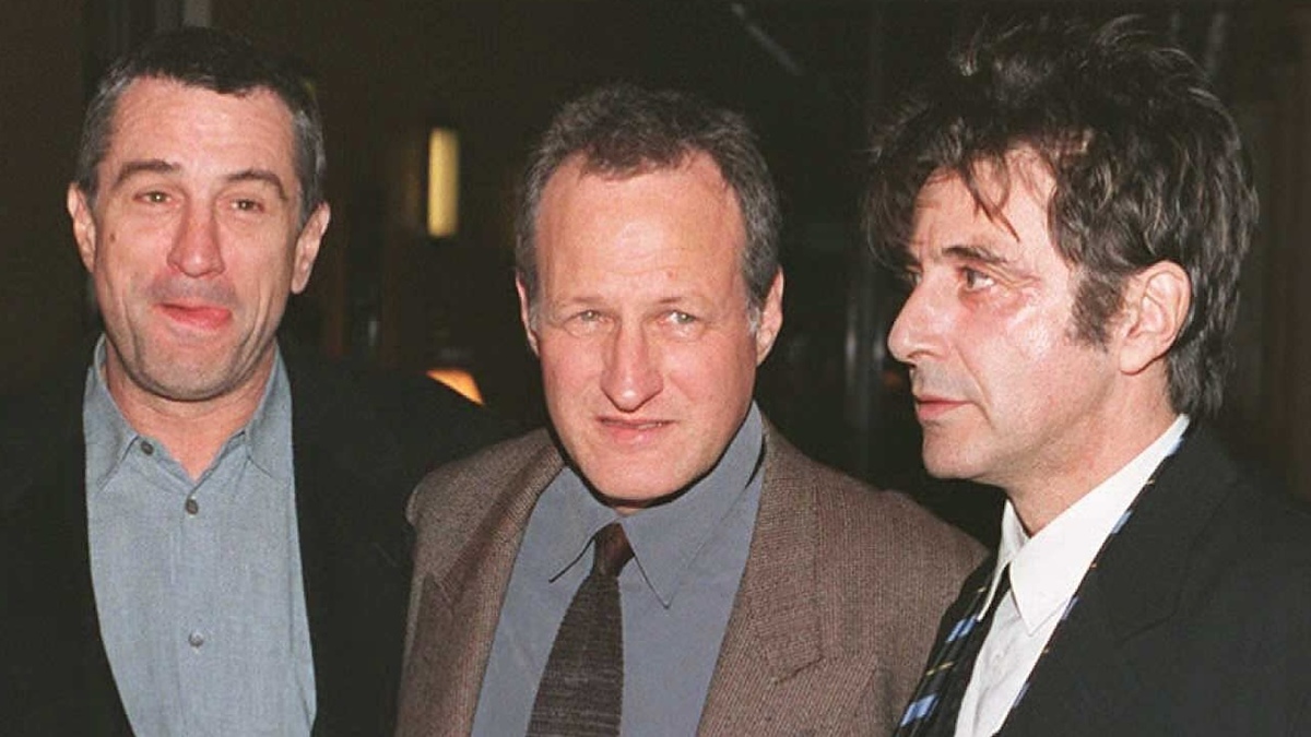Three men are standing together: Robert De Niro, Michael Mann, and Al Pacino. Robert wears a casual shirt, Michael wears a suit, and Al wears a suit with a tie
