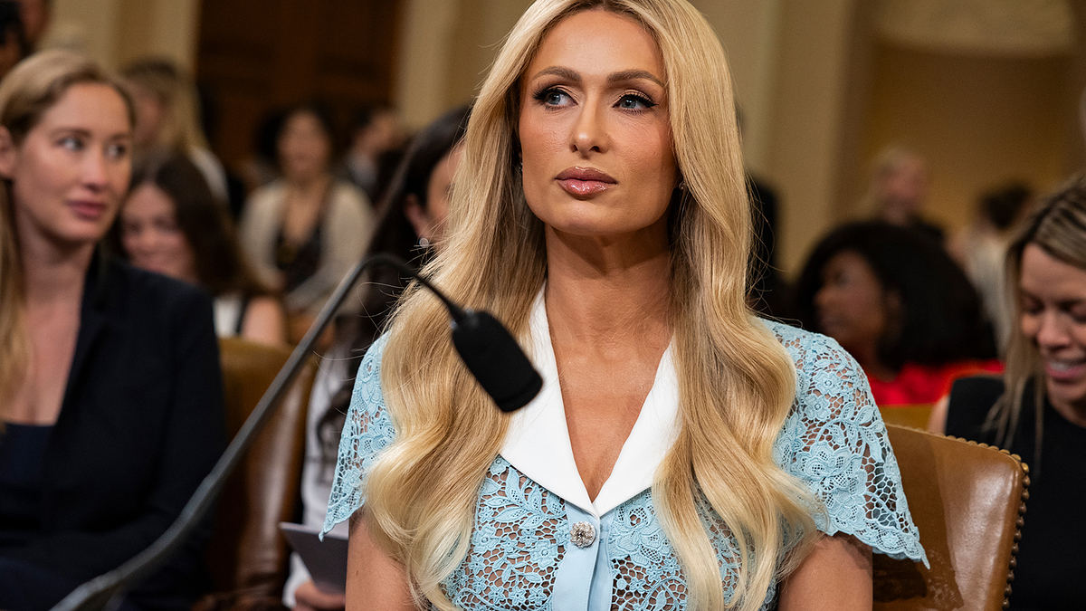 Paris Hilton sits at a hearing, wearing a lace dress with a white collar. Several people are present in the background