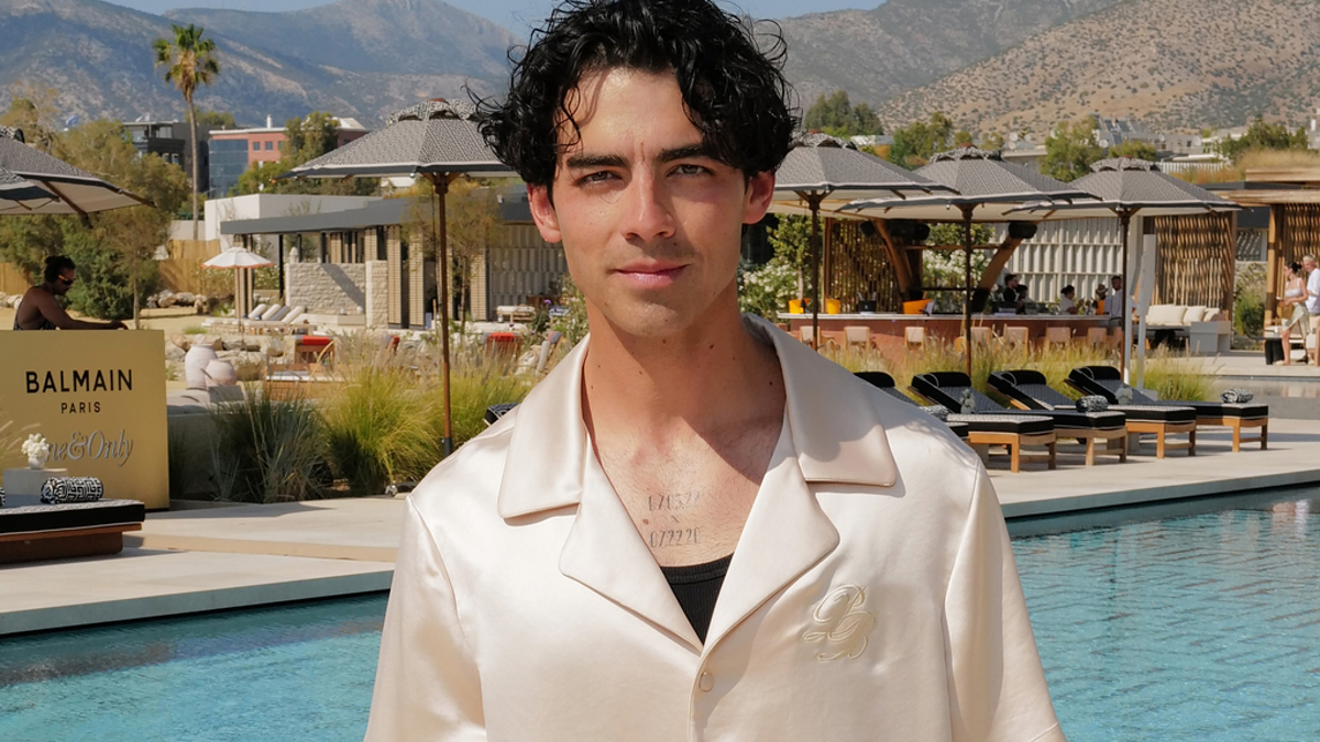 Joe Jonas stands by a poolside wearing a short-sleeved silk shirt and black shorts, with sunglasses in his hand