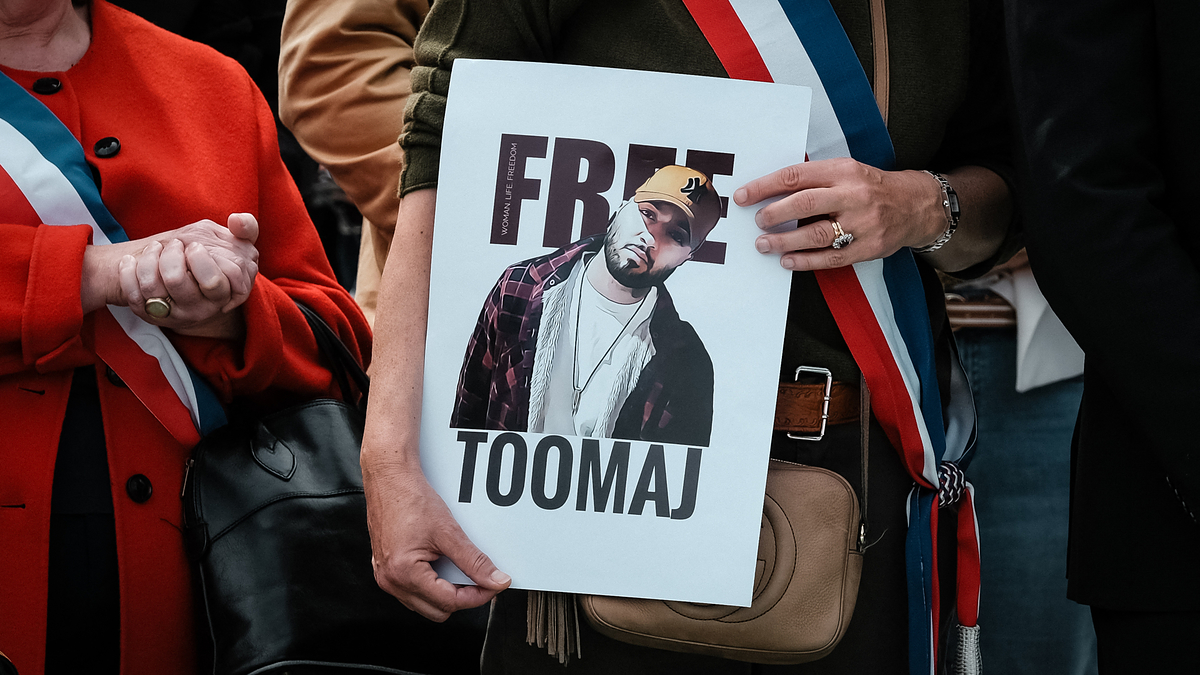 A person holding a "FREE TOOMAJ" sign with an image of Toomaj in a plaid shirt and yellow cap, surrounded by people wearing sashes