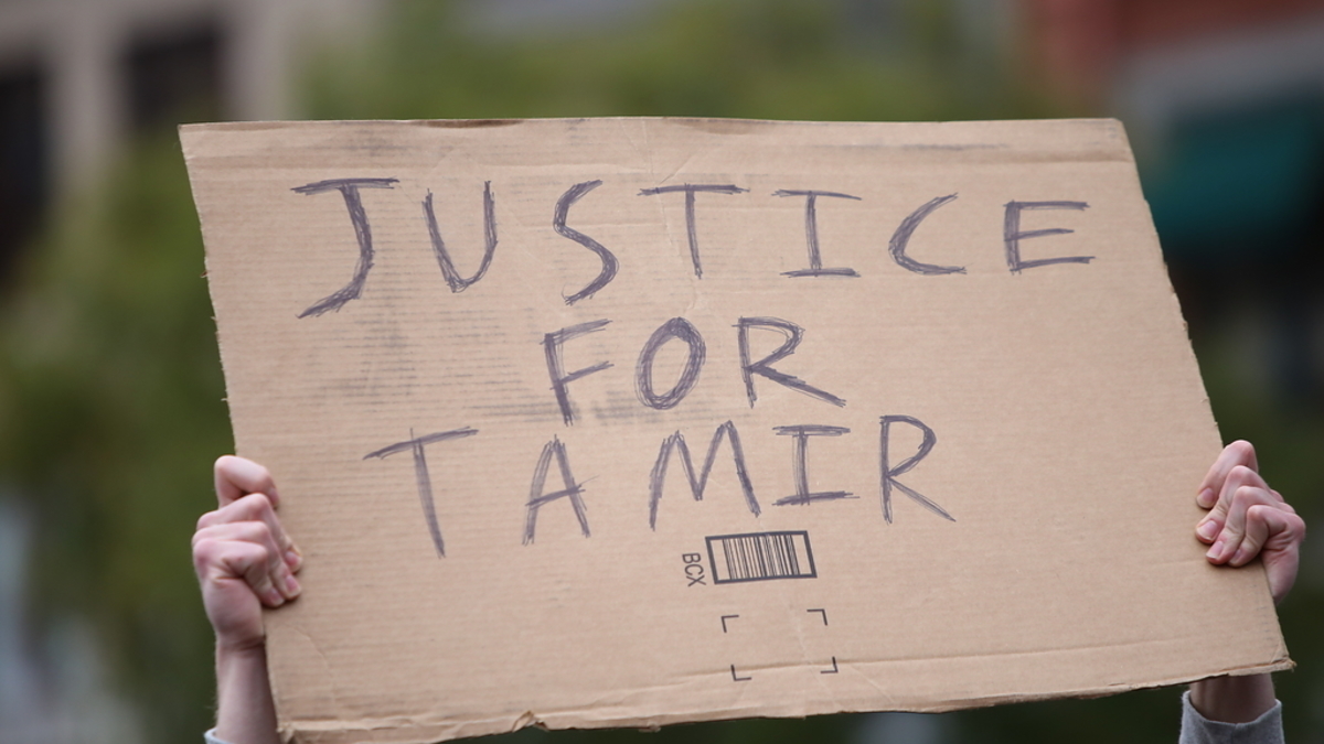 A cardboard sign with "Justice for Tamir" written in bold letters is being held up by two hands, likely at a protest