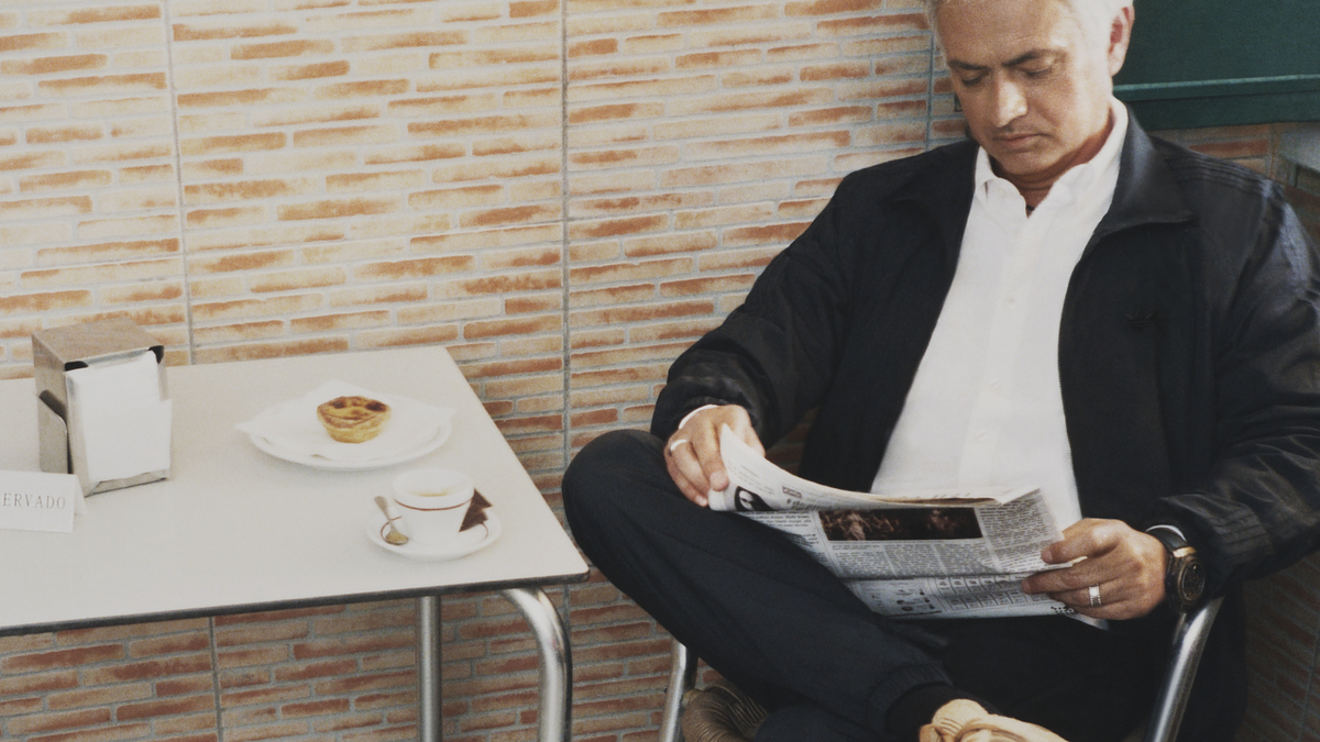 José Mourinho sits at a cafe table reading a newspaper, wearing casual clothing and stylish sneakers