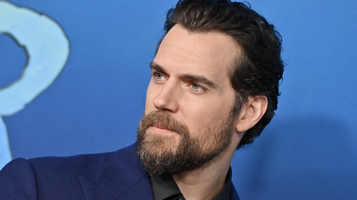 Henry Cavill at an event, facing sideways with a thoughtful expression, wearing a dark suit and black shirt against a blue backdrop