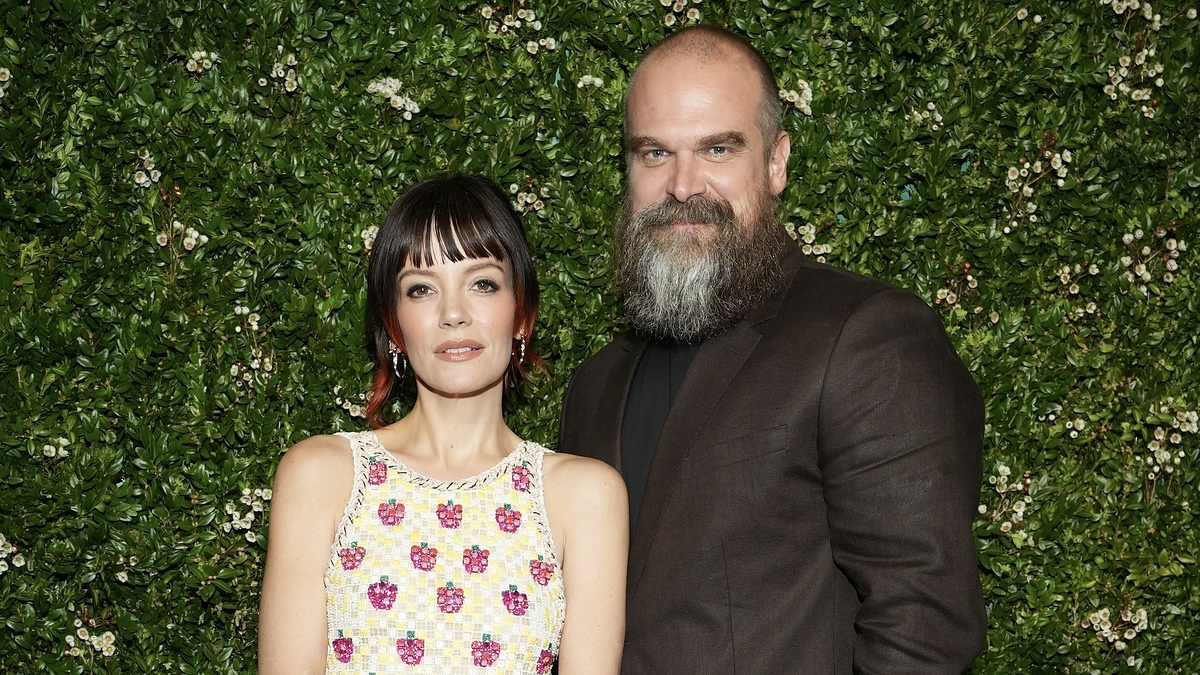 Lily Allen, in a patterned sleeveless dress, and David Harbour, wearing a dark suit, pose together in front of a greenery backdrop