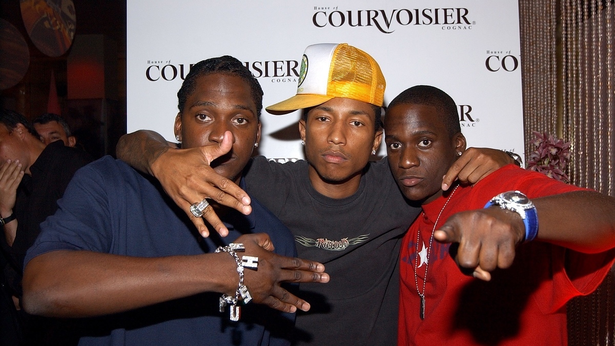 Pusha T, Pharrell Williams, and Malice pose together at a Courvoisier event. Pusha T and Malice point at the camera while Pharrell Williams wears a tilted hat