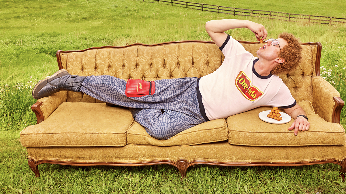 Man in casual clothing lays on an antique sofa in a grassy field, eating snacks from a small plate, with a Hulu logo in the bottom right corner