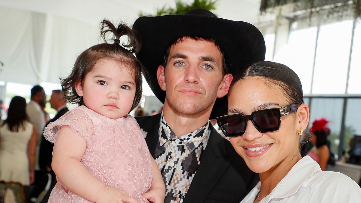Man holding a child in a pink dress while standing next to a woman wearing sunglasses. Names not provided