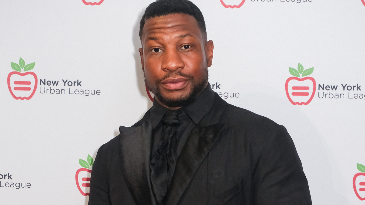 Jonathan Majors in a black suit and tie at the New York Urban League event, standing in front of a step and repeat backdrop with the event logo
