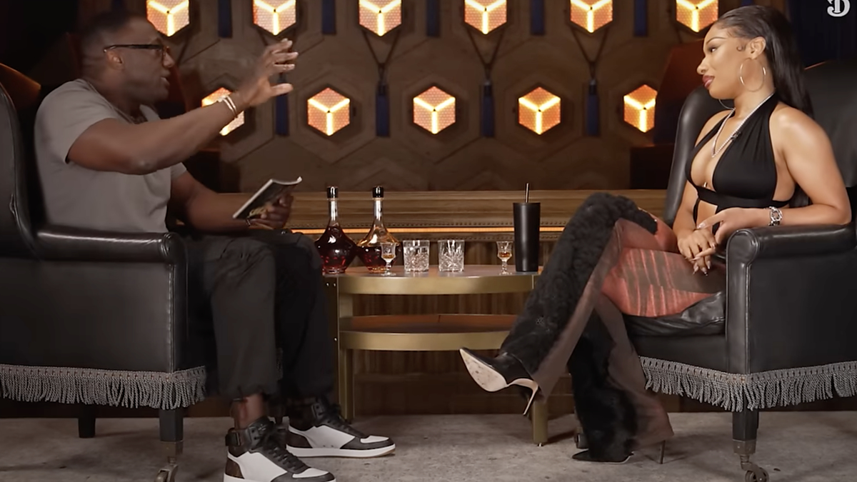 Podcast interview: Megan Thee Stallion in a chic outfit talks with host in casual attire. They sit opposite each other with a table of drinks in between