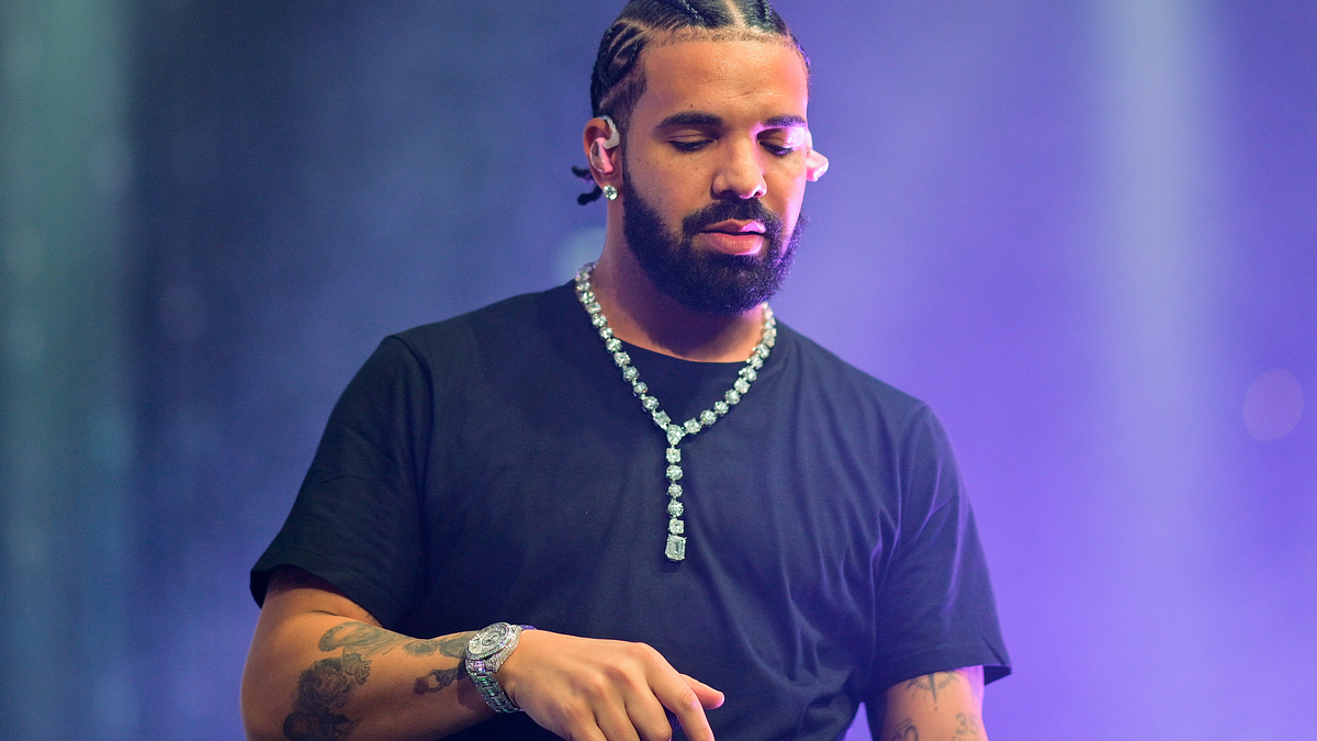 Drake on stage performing, wearing a black T-shirt, diamond necklace, and a watch, with braided hair styled back
