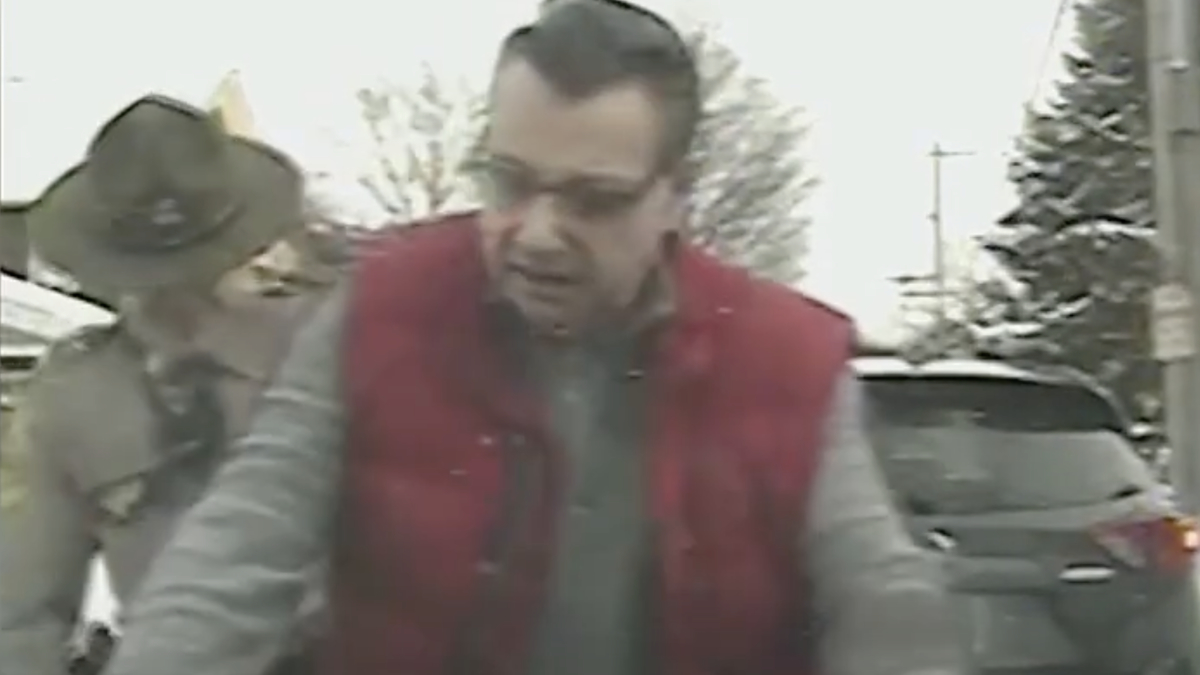 A man wearing a red vest and glasses is being detained by an officer in a uniform and hat, next to a dark car in a snow-covered area