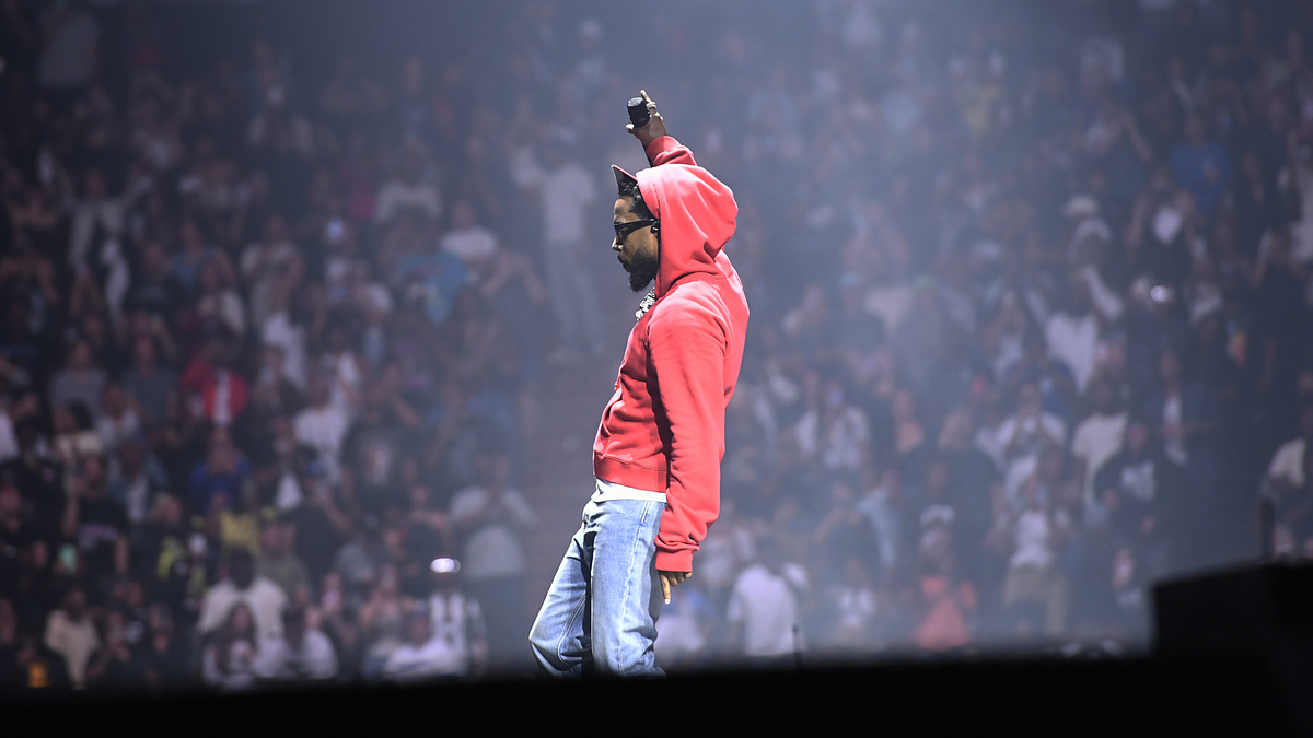 Kendrick Lamar on stage, wearing a hoodie and jeans, raising his right hand with a microphone in front of a large concert audience