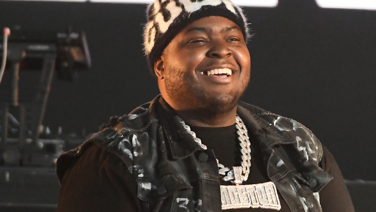 Sean Kingston performs on stage wearing a vest with a large necklace, while DJ Nyce Hitz is seen in the background working the turntables