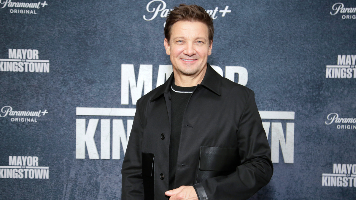 Jeremy Renner poses in a black jacket at the premiere of "Mayor of Kingstown," against a backdrop with Paramount+ branding