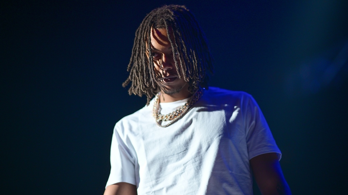 A person with shoulder-length hair wearing a white t-shirt and multiple gold chains around their neck stands under a spotlight on stage